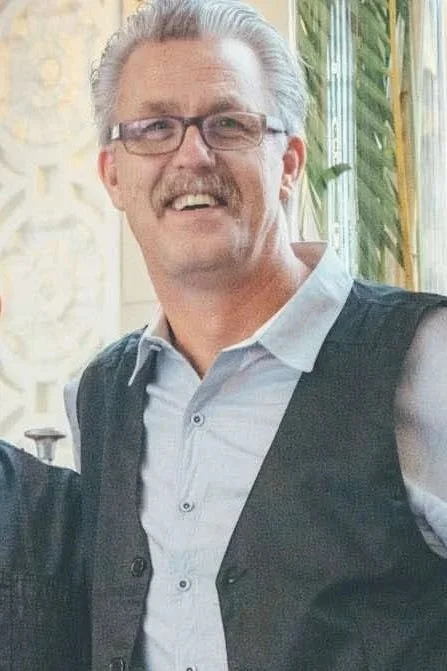 A smiling man with gray hair, glasses, and a mustache, wearing a light blue shirt and a dark vest, standing indoors near a plant and a window.