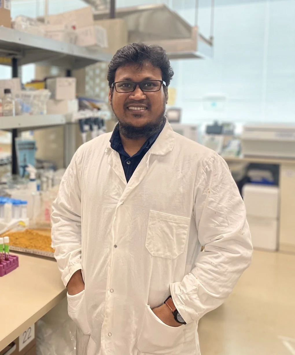 A man in a white lab coat, standing in front of a laboratory with shelves of bottles and scientific equipment.