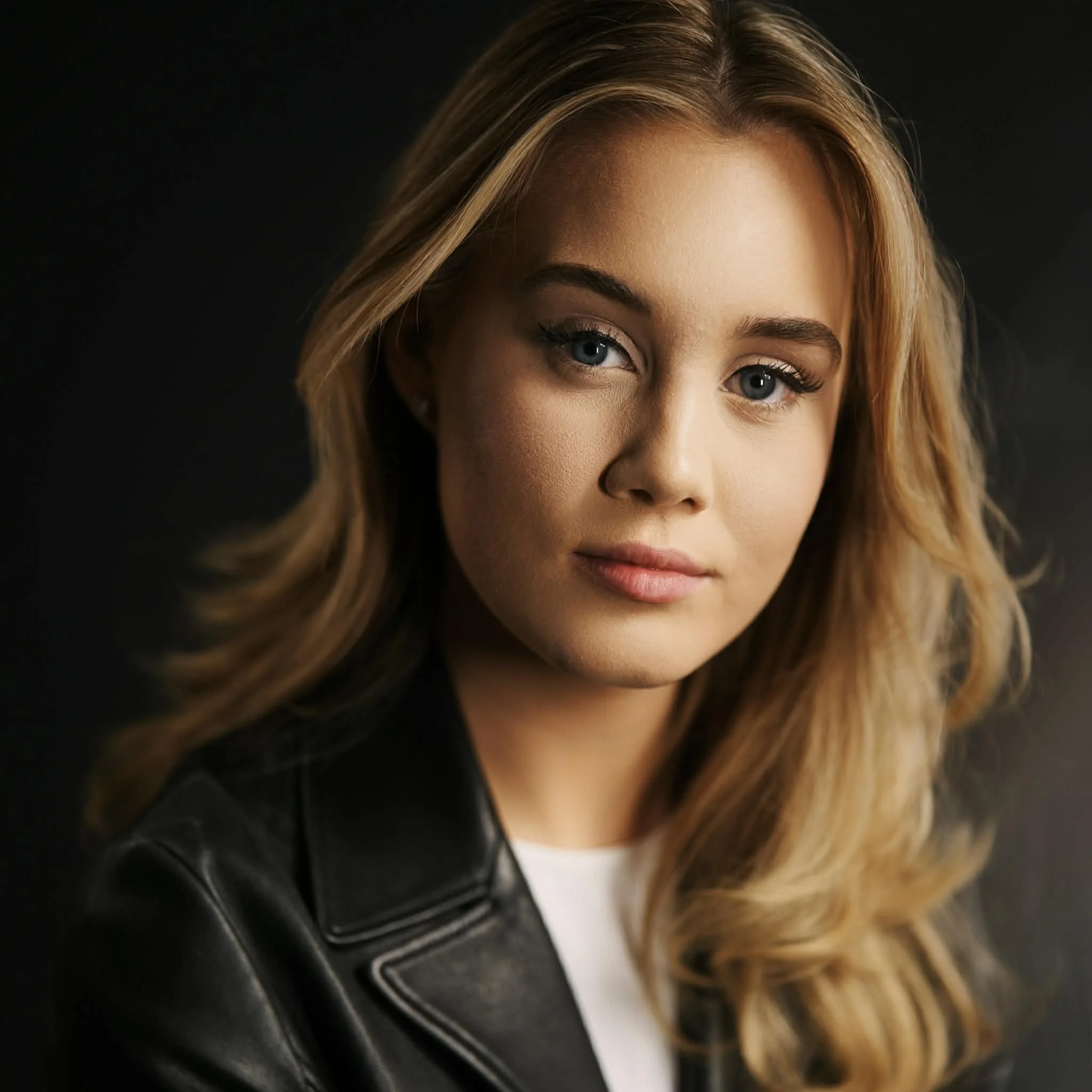A close-up portrait of a young woman with wavy blonde hair, blue eyes, light makeup, wearing a black leather jacket and a white top against a dark background.