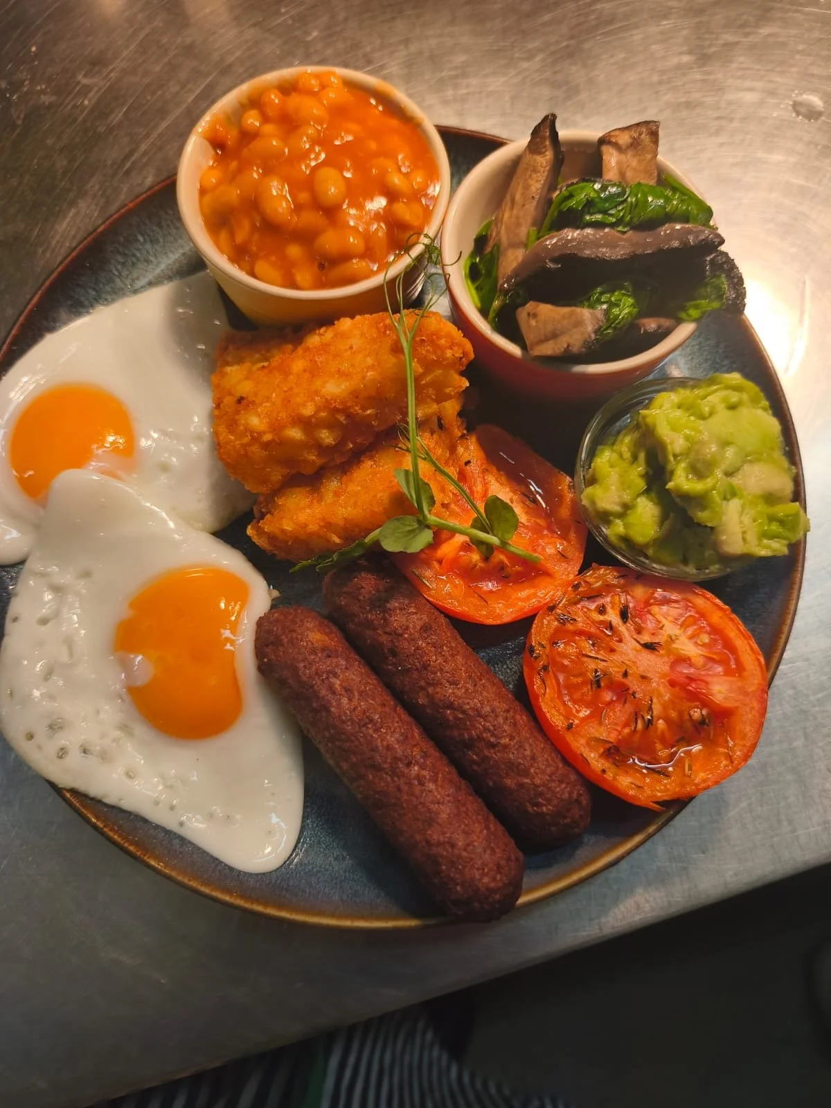 Full English breakfast with fried eggs, sausages, baked tomato, guacamole, roasted vegetables, baked beans, and fried chicken on a decorative plate.