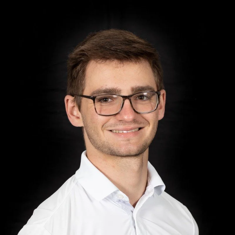 A young man with brown hair, glasses, and a light complexion, smiling and wearing a white collared shirt, against a black background.