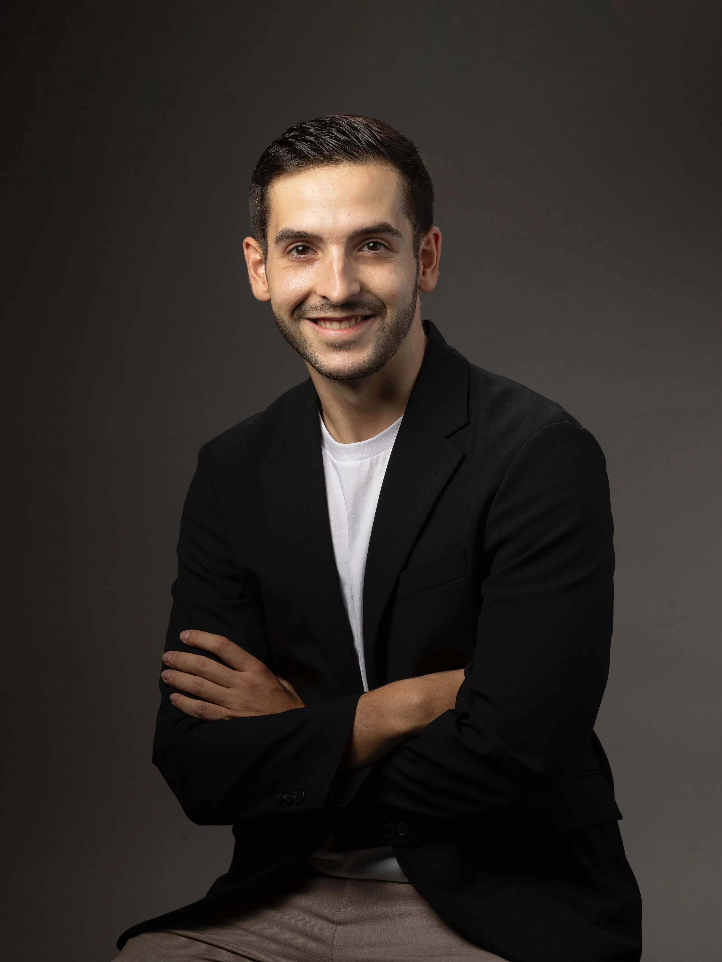 A young man with dark hair, dressed in a black blazer and white shirt, smiling with arms crossed against a dark grey gradient background.