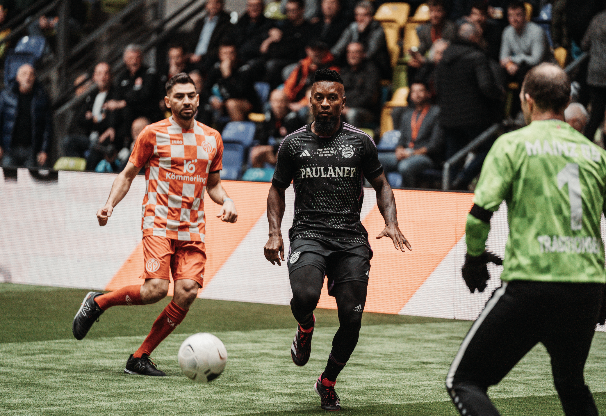 Soccer players in action on a green field during a match, with a crowd of spectators in the background. One player in an orange and white checkered jersey and another in a black jersey are running toward the ball, while the goalkeeper in a green jersey with the number 1 is preparing to defend.