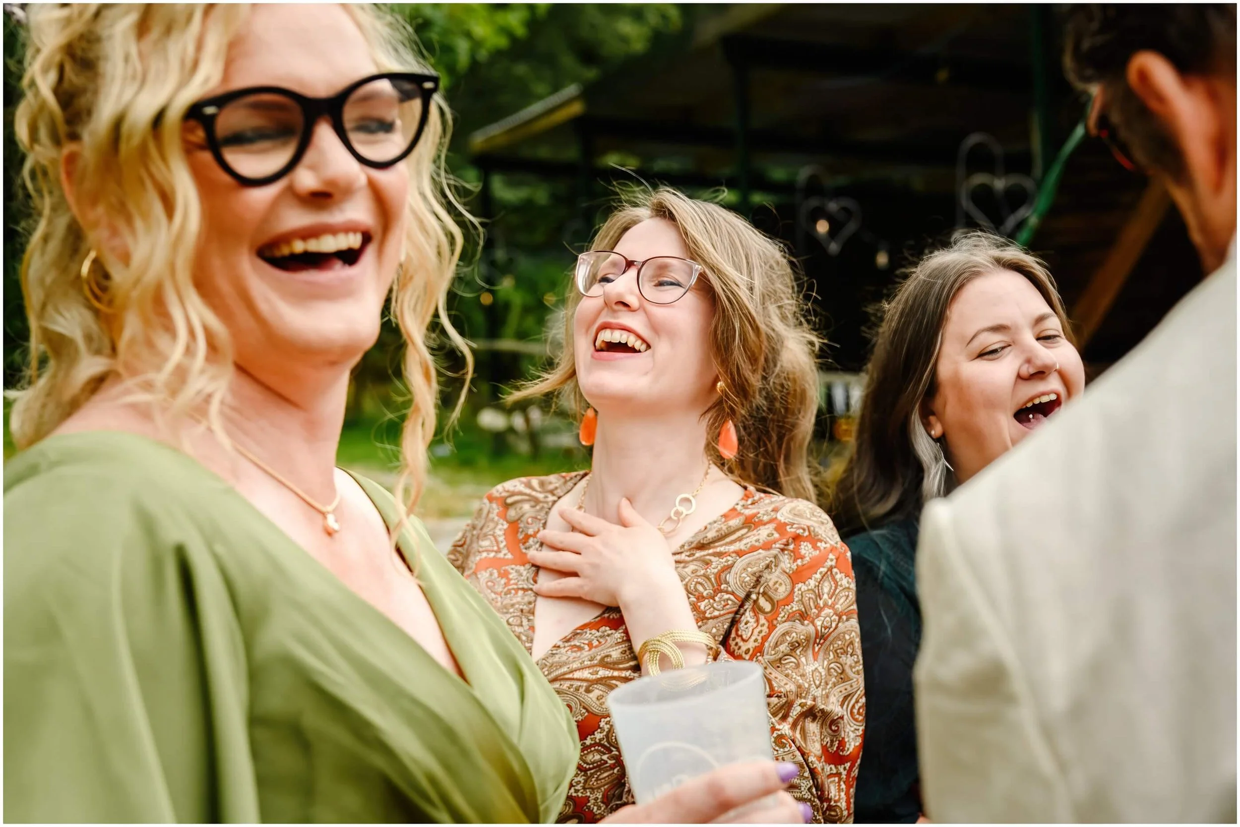 A group of women laughing and smiling at an outdoor gathering. One woman is wearing black glasses and a green dress, another is wearing clear glasses and an orange paisley dress, and a third woman has long dark hair and a nose ring.