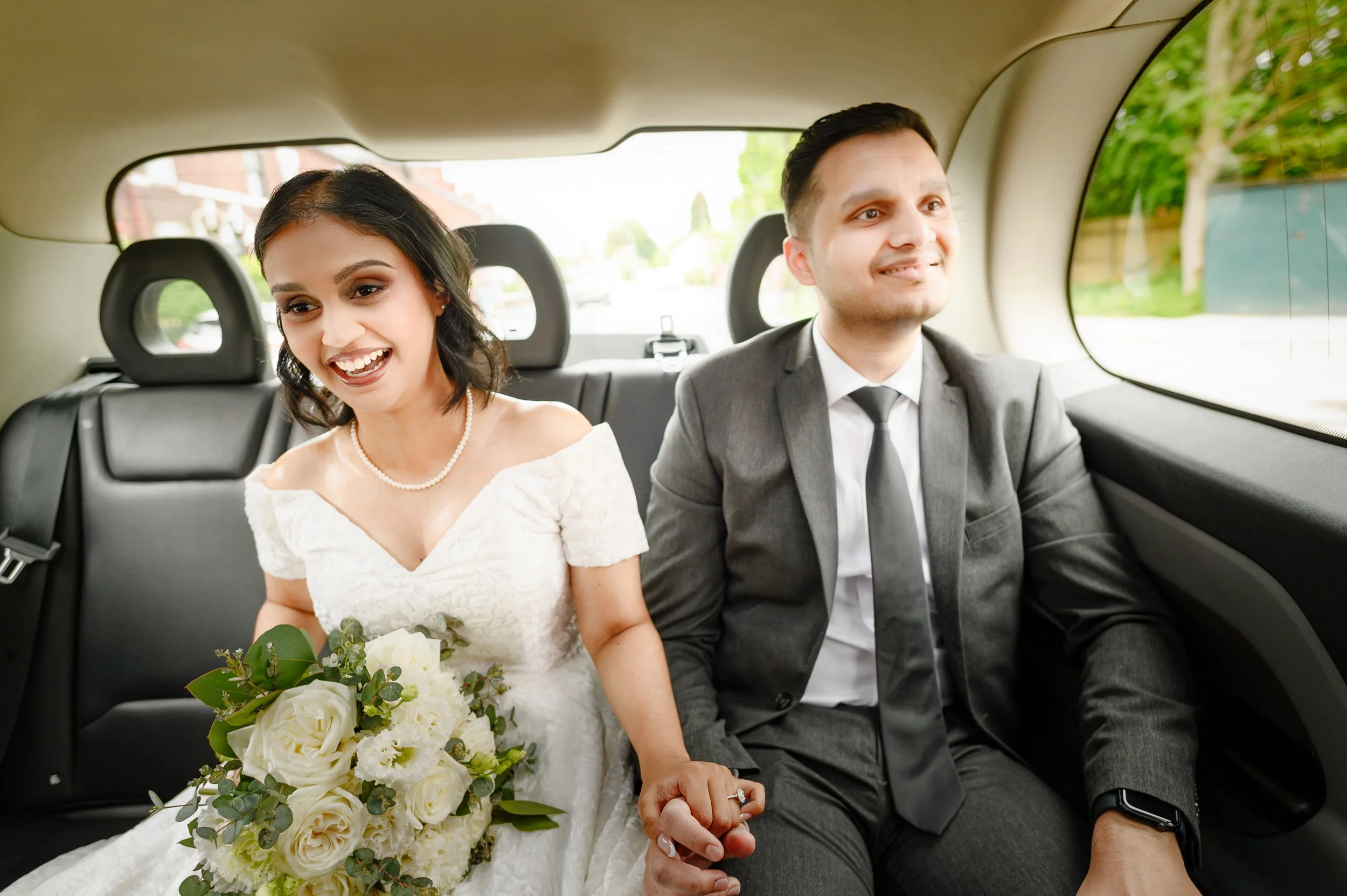 A newlywed couple in wedding attire sitting in the back of a car, holding hands, with the bride holding a bouquet of white roses and green foliage.