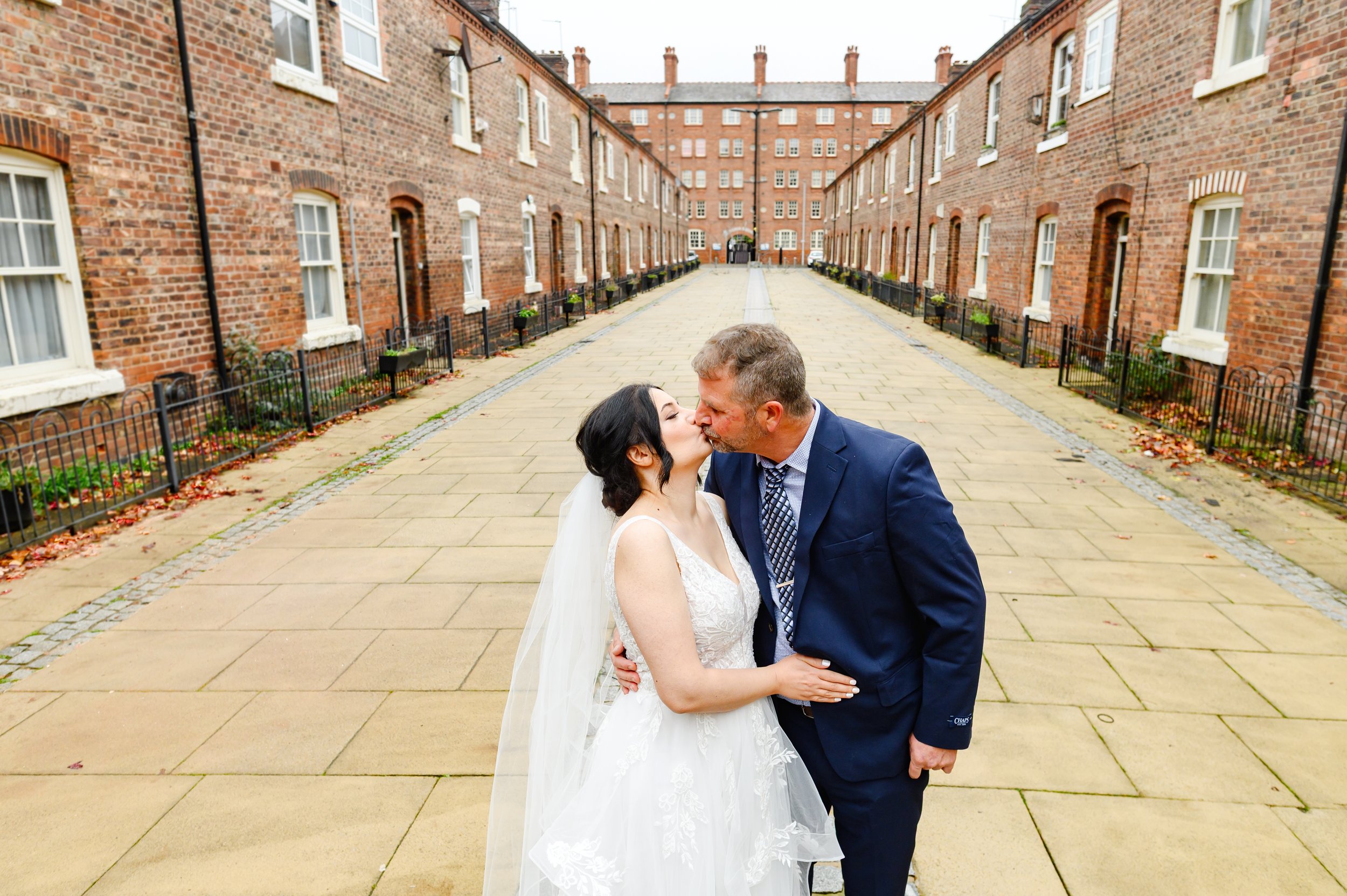 A bride and groom sharing a kiss outdoors in a courtyard surrounded by red brick buildings with multiple windows and small garden beds.