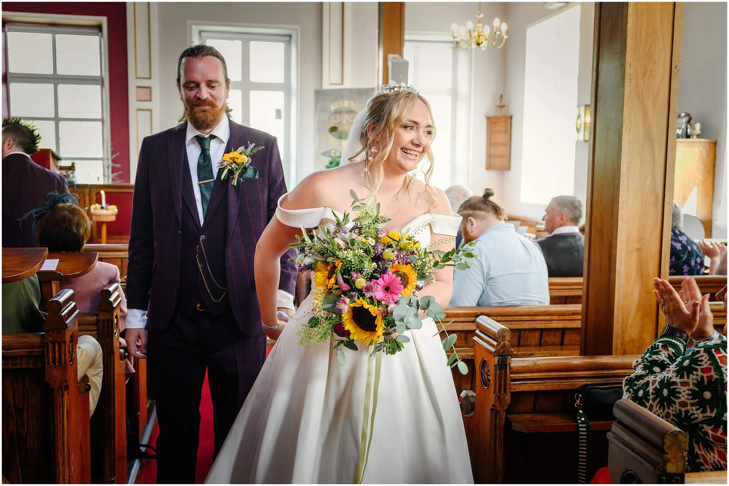 A bride in a white wedding dress carrying a colorful bouquet walking down the aisle with a man in a suit behind her in a church.