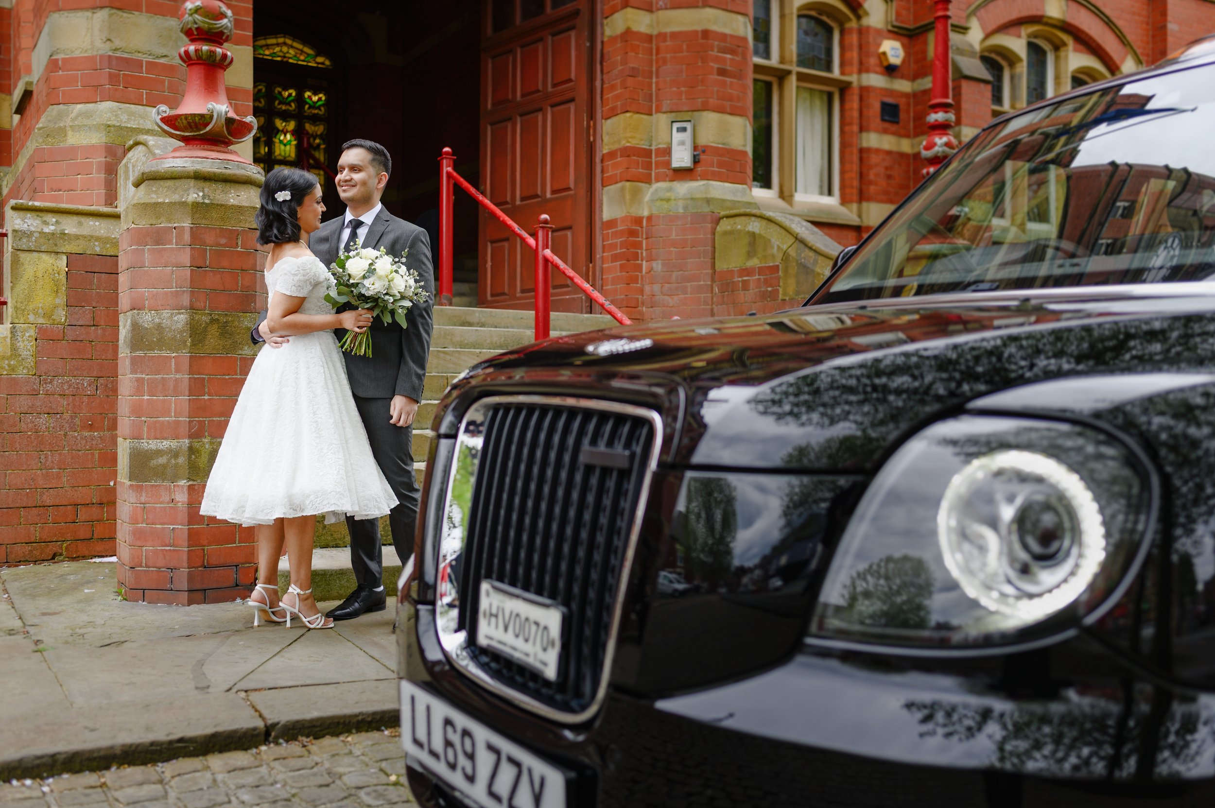 A newlywed couple stands outside a red-brick church, smiling and gazing at each other. The bride is wearing a white dress and holding a bouquet, and the groom is in a gray suit. A black luxury car with a visible license plate is parked nearby.