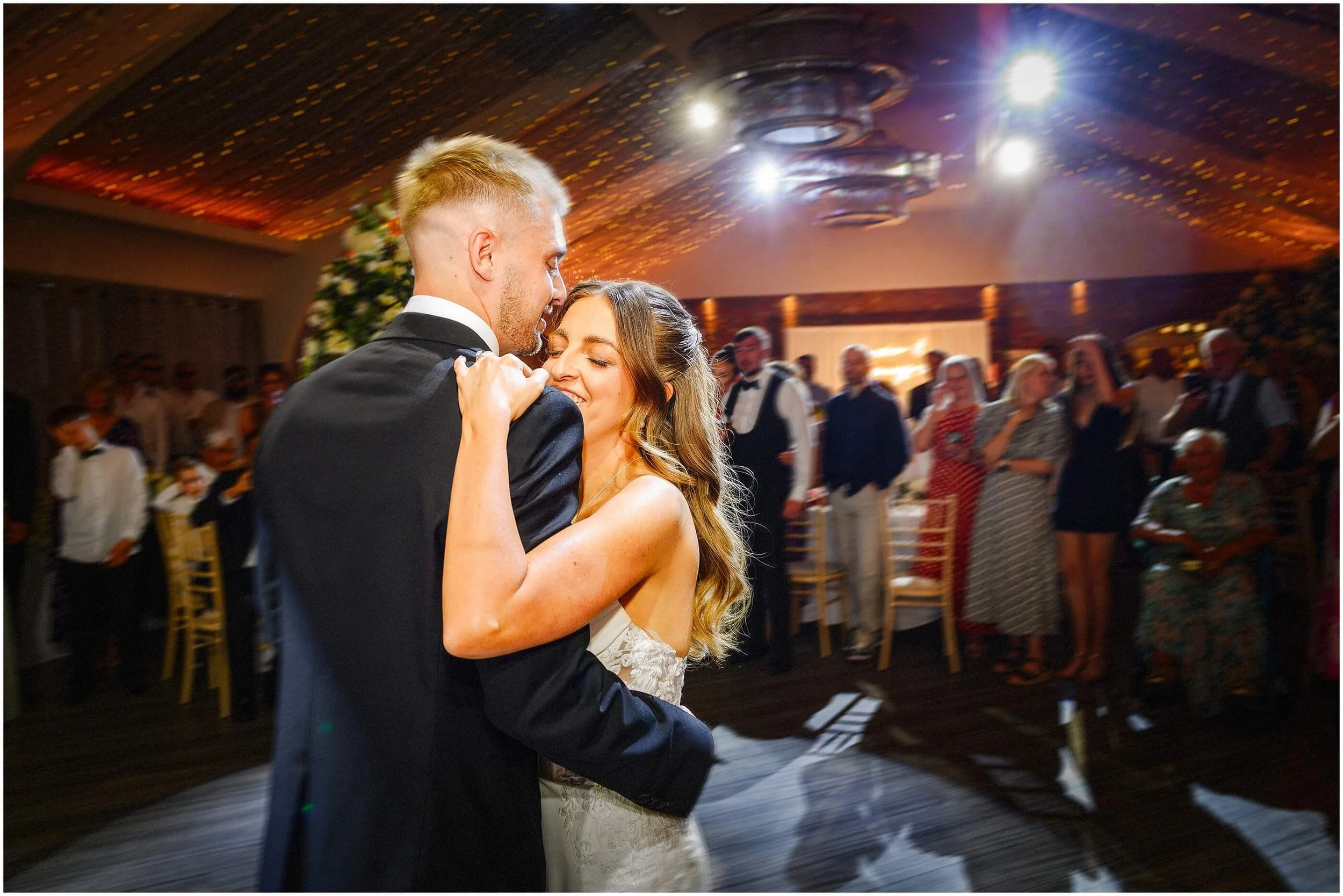 Couple dancing closely, smiling, during a celebration in a decorated indoor venue with wedding guests watching.