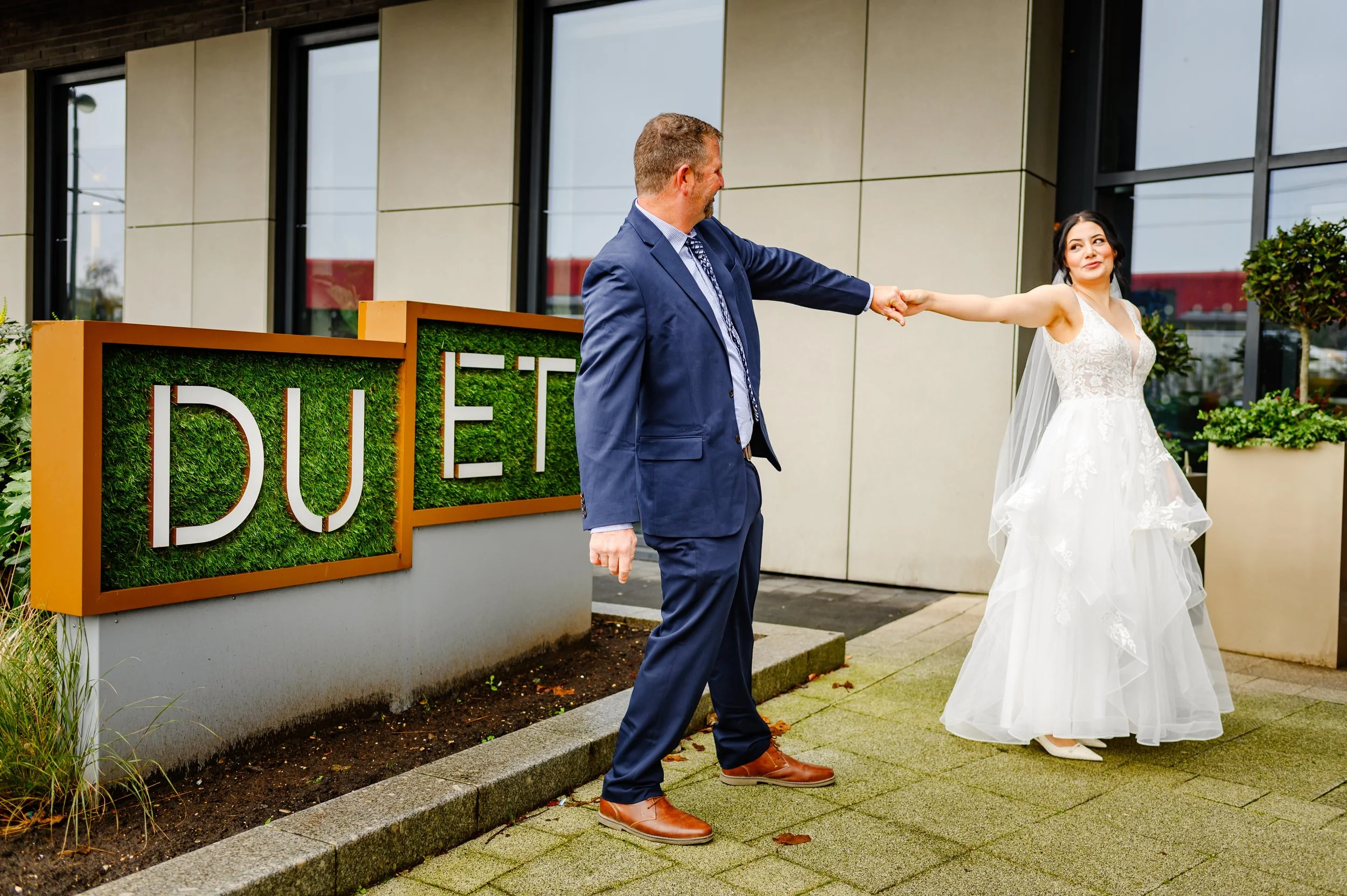 A bride and groom holding hands outside a modern building during a wedding ceremony, with a sign that reads 'DUE' partially visible.