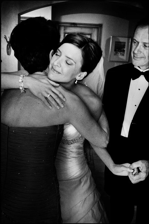 Bride hugging a woman in an embrace, with a man in a tuxedo standing nearby, in a room with framed artwork on the wall.