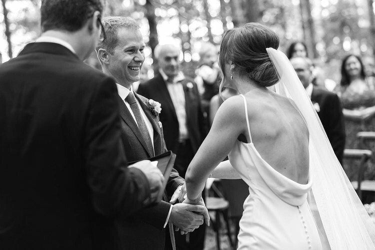 A wedding ceremony outdoors with a bride and groom holding hands, smiling at each other, with an officiant and guests in the background.