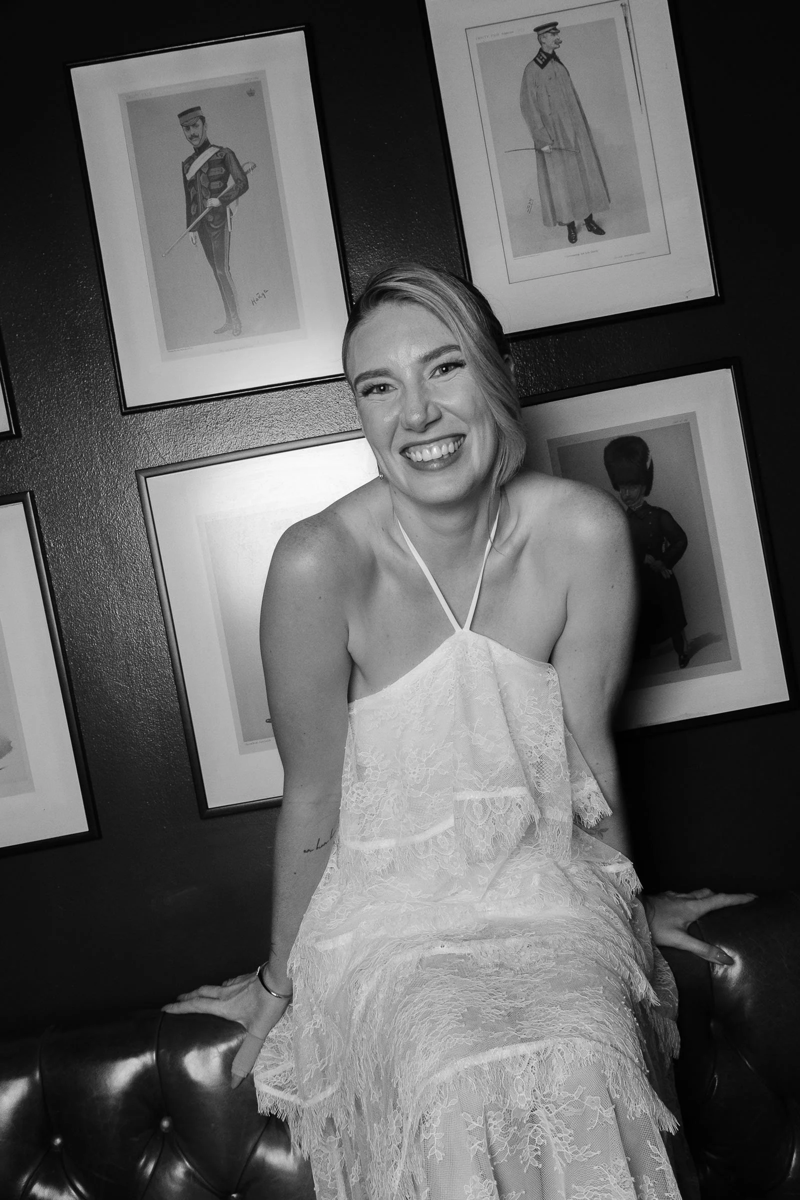 A smiling woman in a white lace dress sitting on a leather couch with framed artwork of military figures on a dark wall behind her.