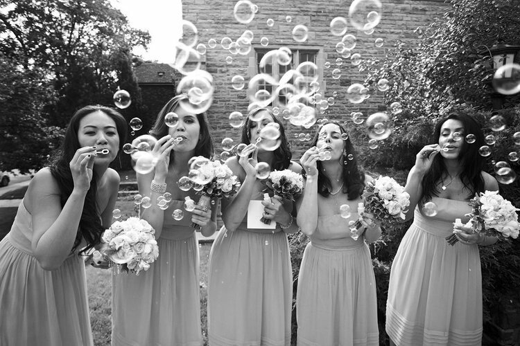 Five women in matching dresses blowing bubbles and holding bouquets, standing outside near a building and trees.
