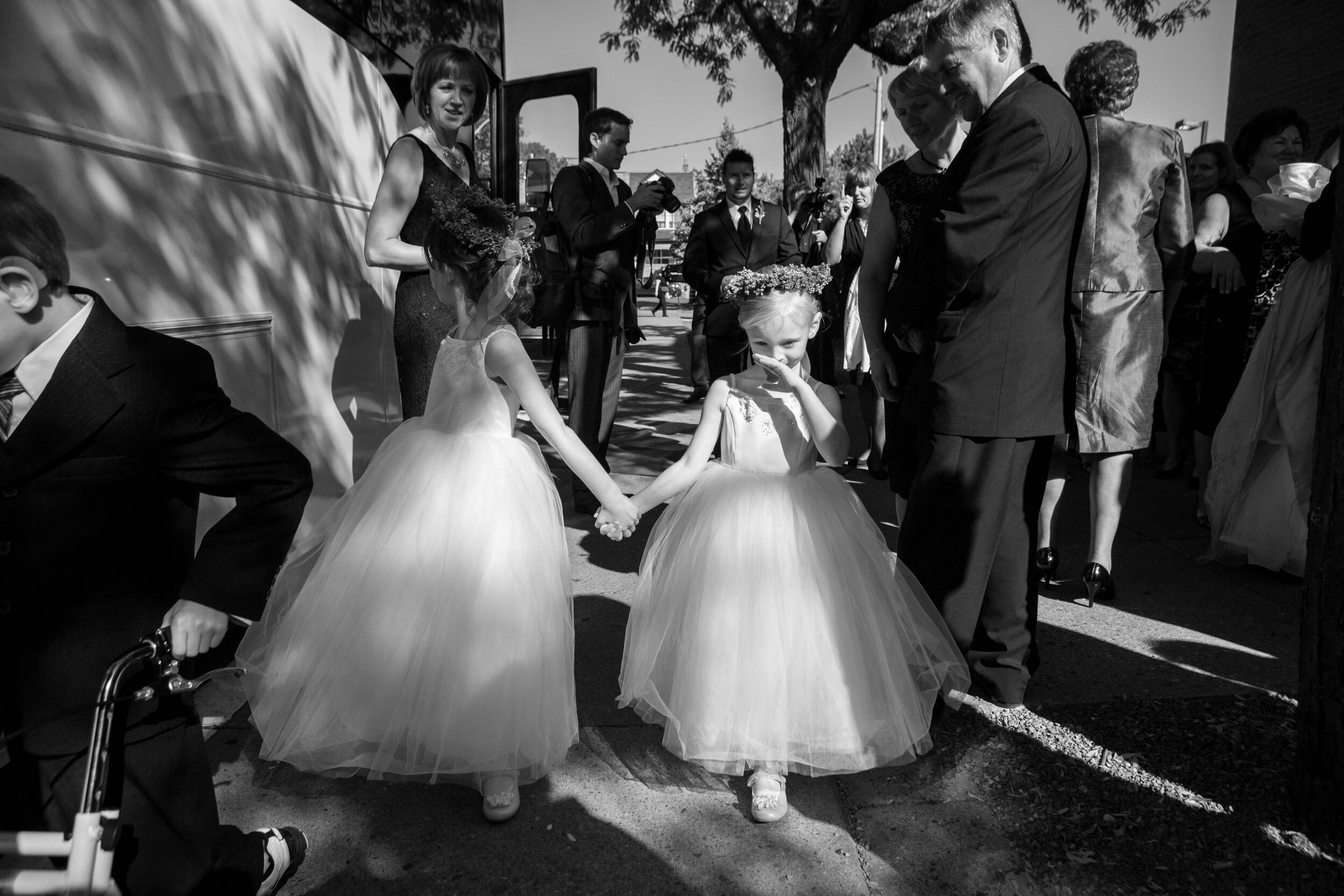 Two young girls in white dresses holding hands and walking outdoors during a wedding celebration, with adults and photographers in the background.