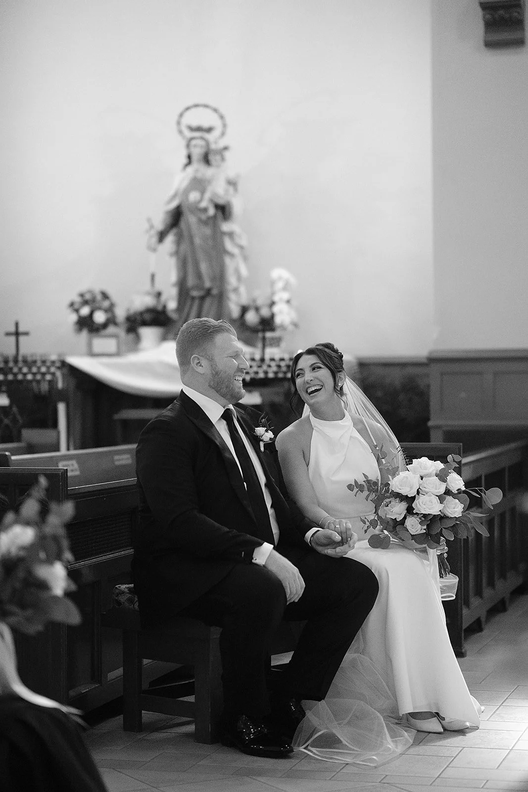 A bride and groom sitting together in a church during their wedding ceremony, smiling and laughing. The bride holds a bouquet of flowers, and there is a religious statue in the background.