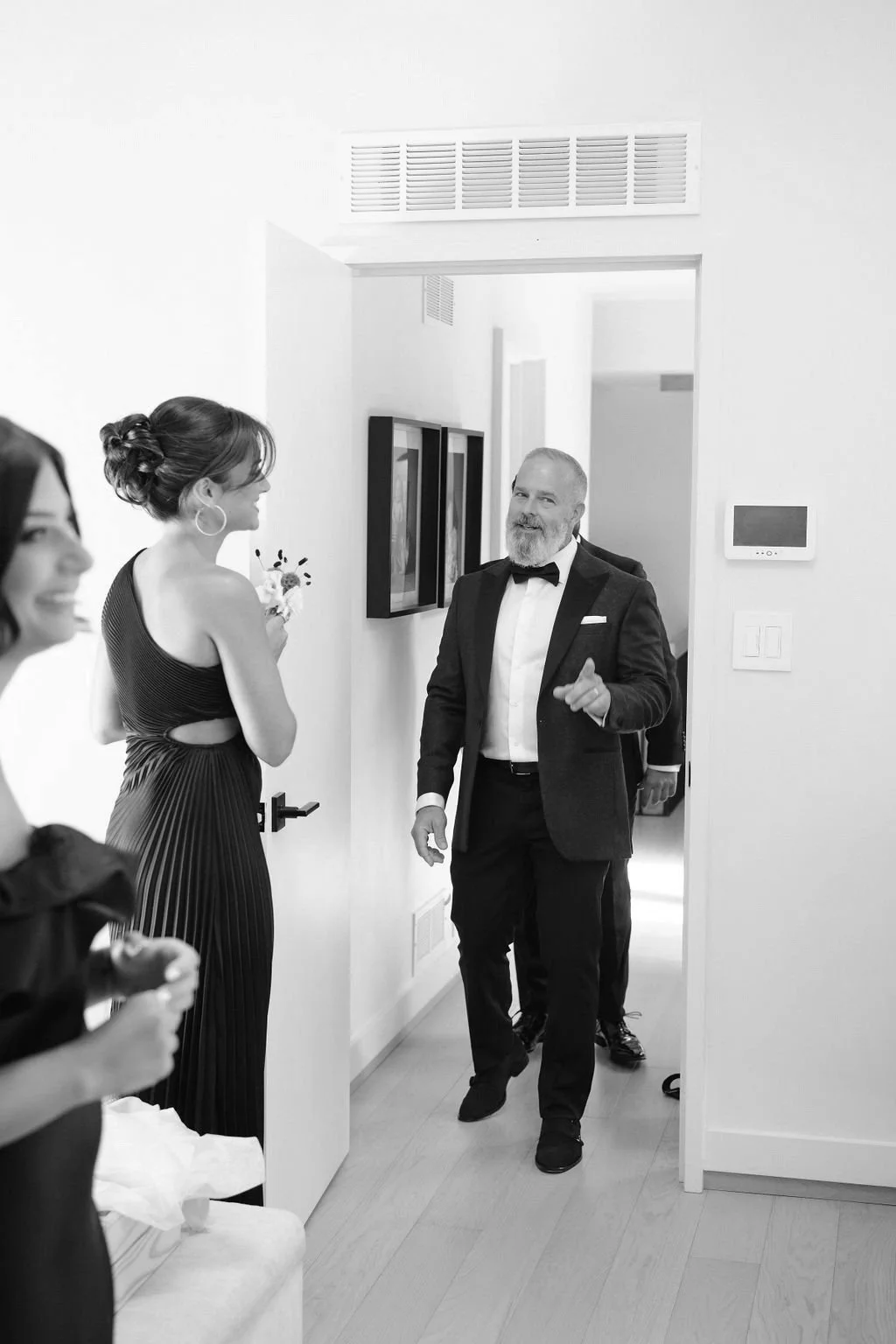 A group of people dressed formally, with a man in a tuxedo smiling and walking into the room while others look on, in a well-lit modern home interior.
