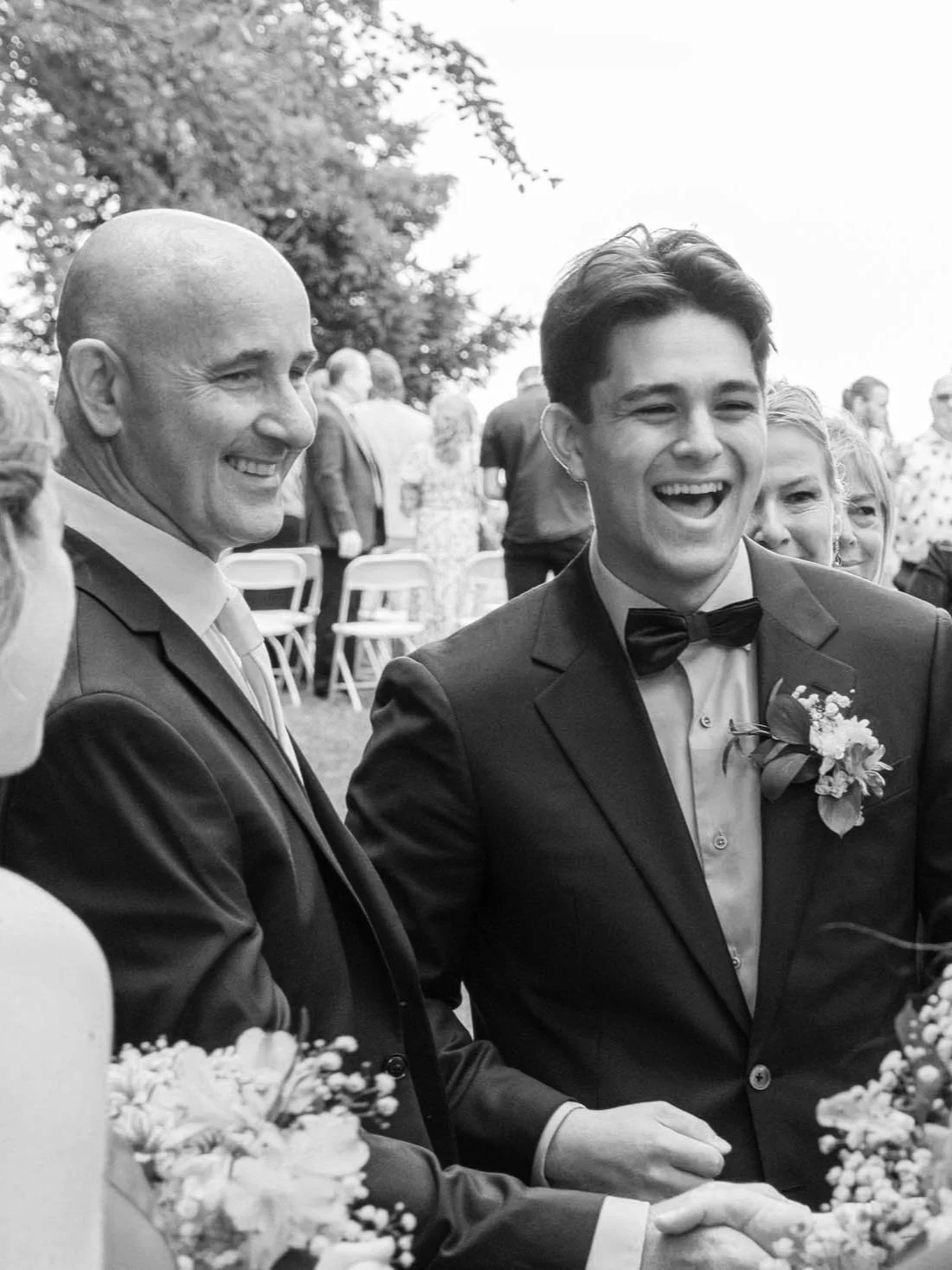 A black and white photo of two men in formal suits, smiling and laughing at an outdoor event. They are surrounded by people and flowers.