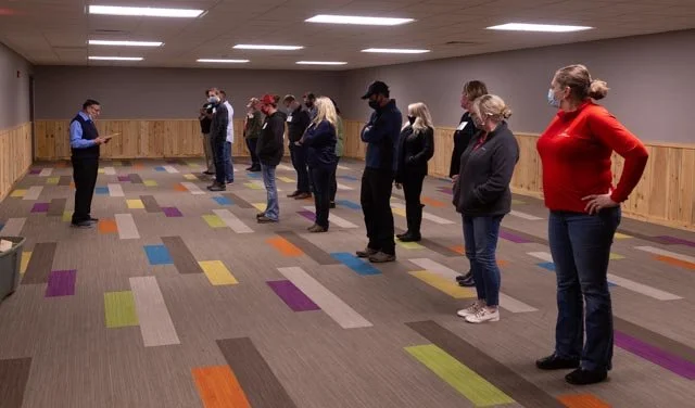 People standing in a line wearing masks, maintaining social distance, indoor setting with colorful patterned carpet.