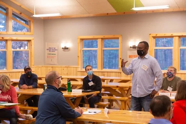 Man wearing glasses and a black mask speaking to a seated group in a room with wooden benches and large windows, all wearing masks.