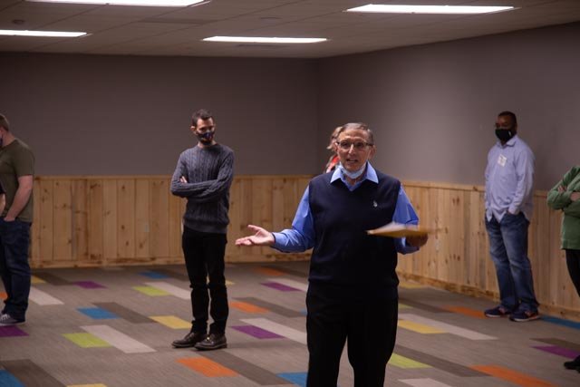 A woman in a blue shirt, dark vest, and face mask speaking to a group of people in a room with a colorful carpet and wooden paneling on the wall.