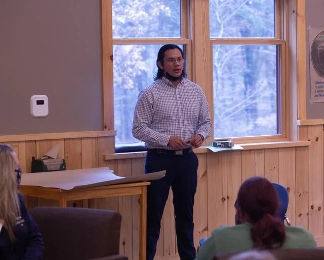 Man speaking to a group of people in a room with wooden walls and large windows.