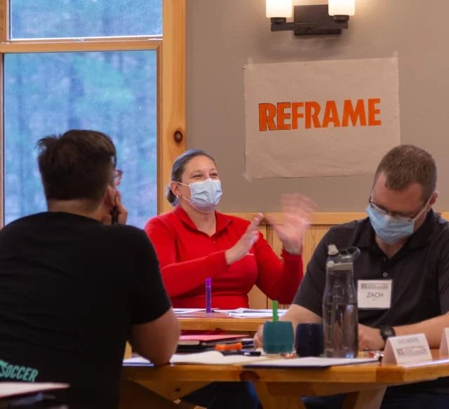 Three people sitting at a table during a discussion, two men and a woman, all wearing face masks; a large paper on the wall reads 'REFRAME' in orange letters.