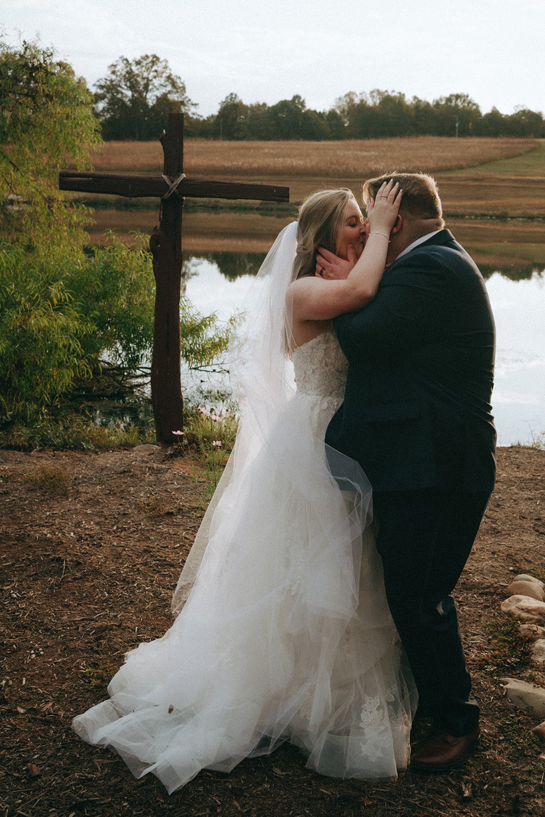 A bride and groom sharing a kiss outdoors near a lake, with a rustic wooden cross in the background.