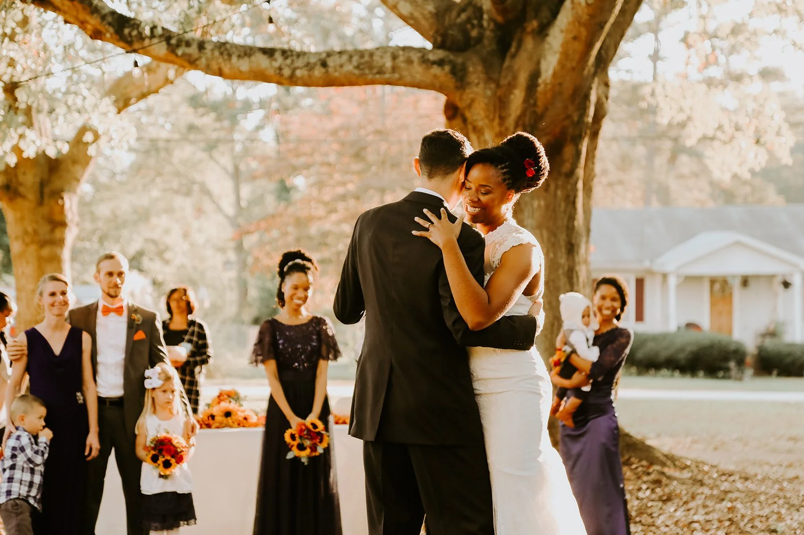 A wedding couple dancing under a large tree, with guests watching and smiling in the background.