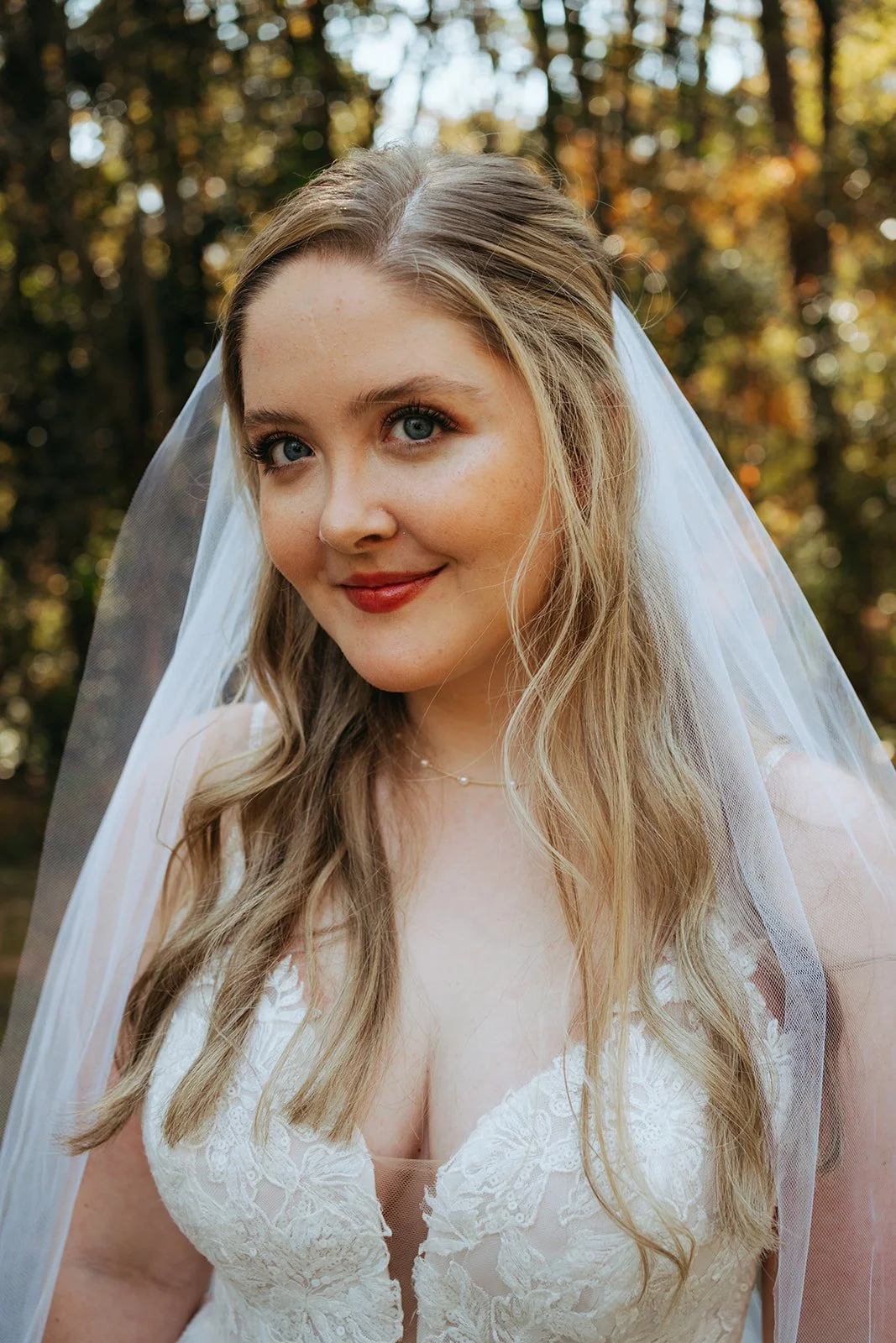 Close-up of a smiling bride with blue eyes and blonde hair, wearing a white lace wedding dress and veil outdoors during autumn.