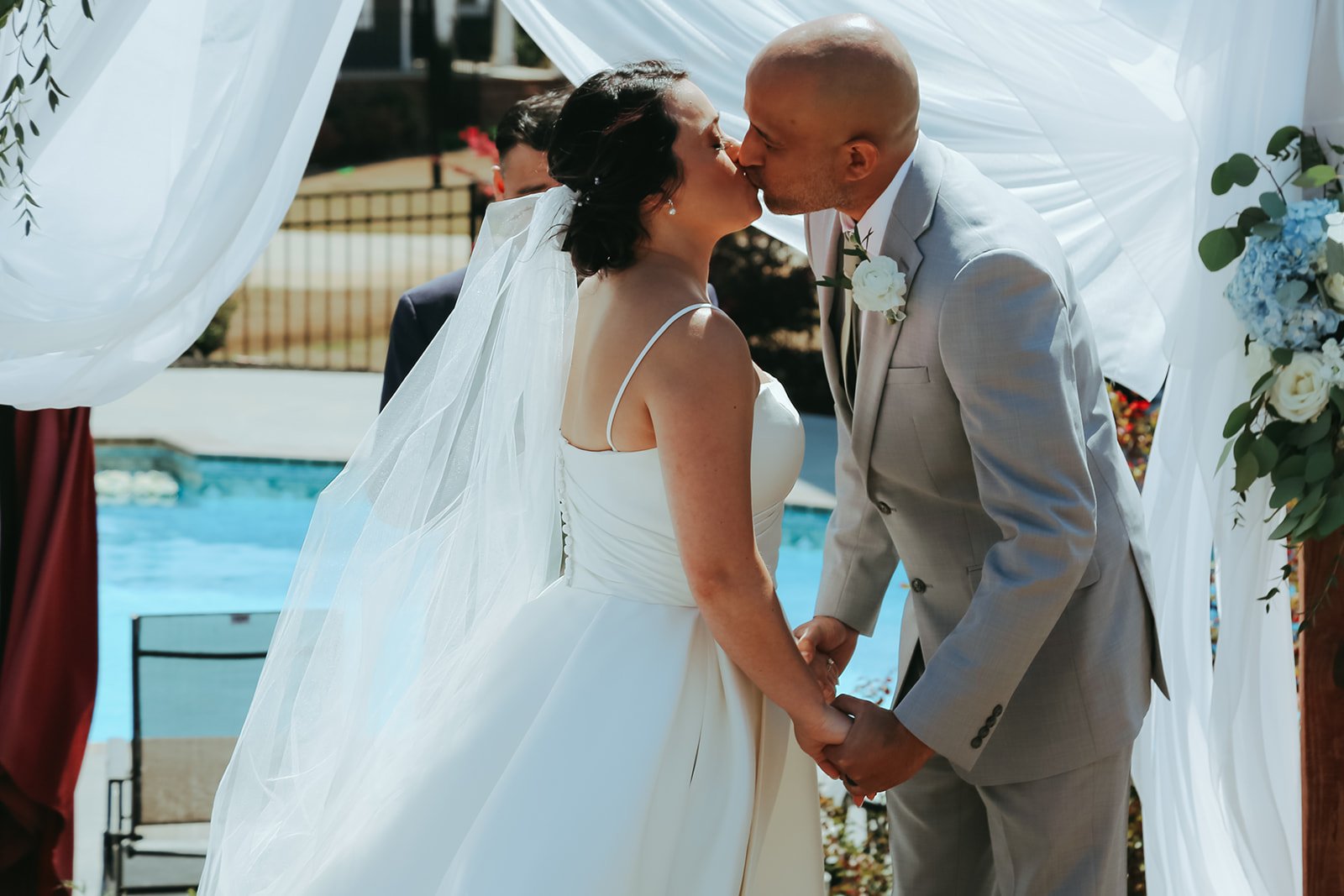 A newlywed couple shares a kiss during their outdoor wedding ceremony by a pool, dressed in wedding attire with a white veil and bouquet, holding hands underneath a white fabric canopy with floral decorations.