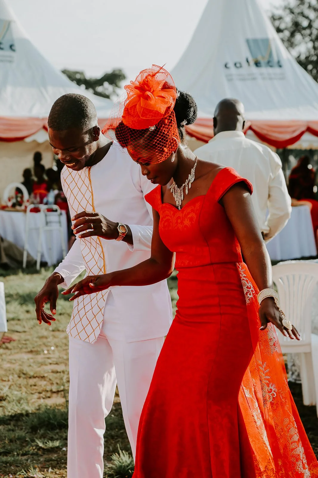 A couple dressed in formal attire celebrating at an outdoor event, with tents and tables in the background.