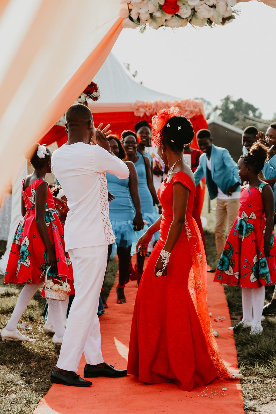A wedding scene with a bride in a red dress and a groom in white, surrounded by friends and family in colorful dresses and suits, under a decorated canopy.
