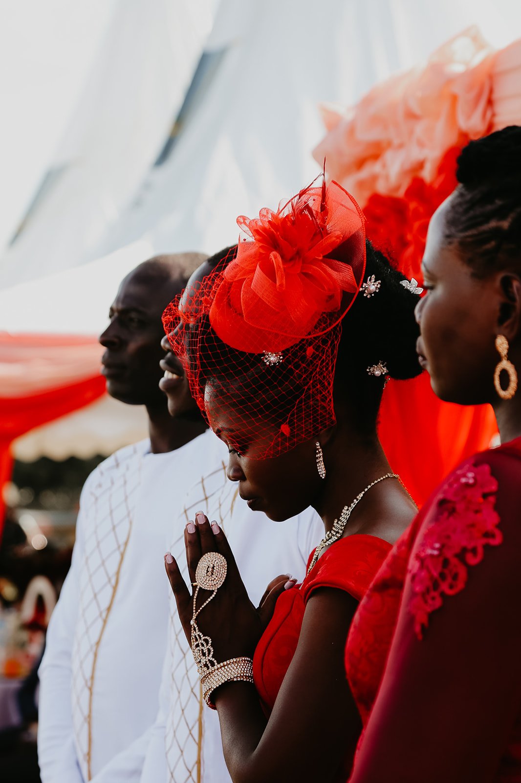 Four people, dressed in formal or traditional clothing, appear to be engaged in a prayer or moment of reflection, with their hands clasped, during an outdoor event under a decorated canopy.