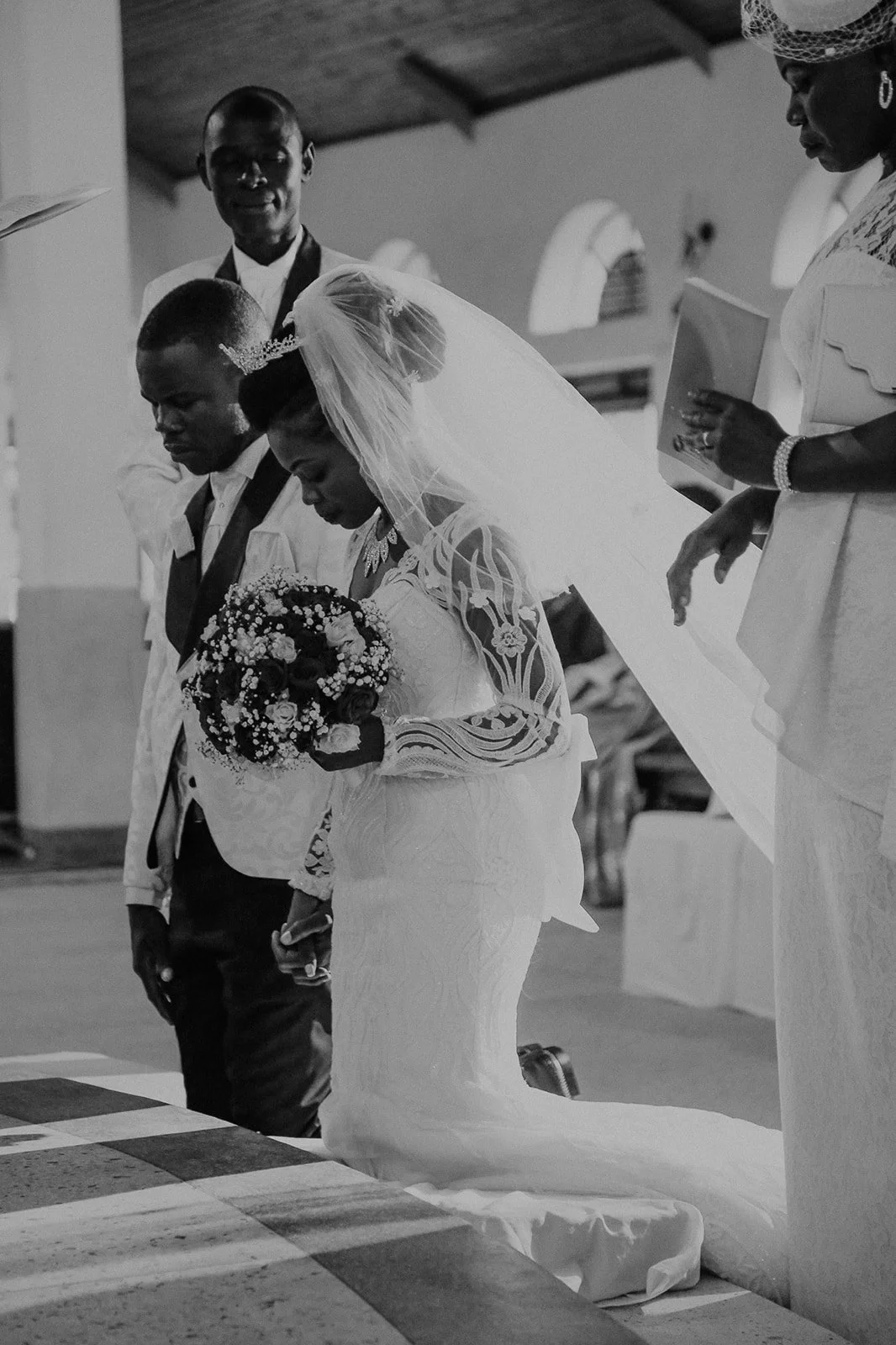 A bride and groom kneel during a wedding ceremony inside a church, surrounded by their wedding party, with the bride holding a bouquet of flowers and wearing a lace dress and veil.