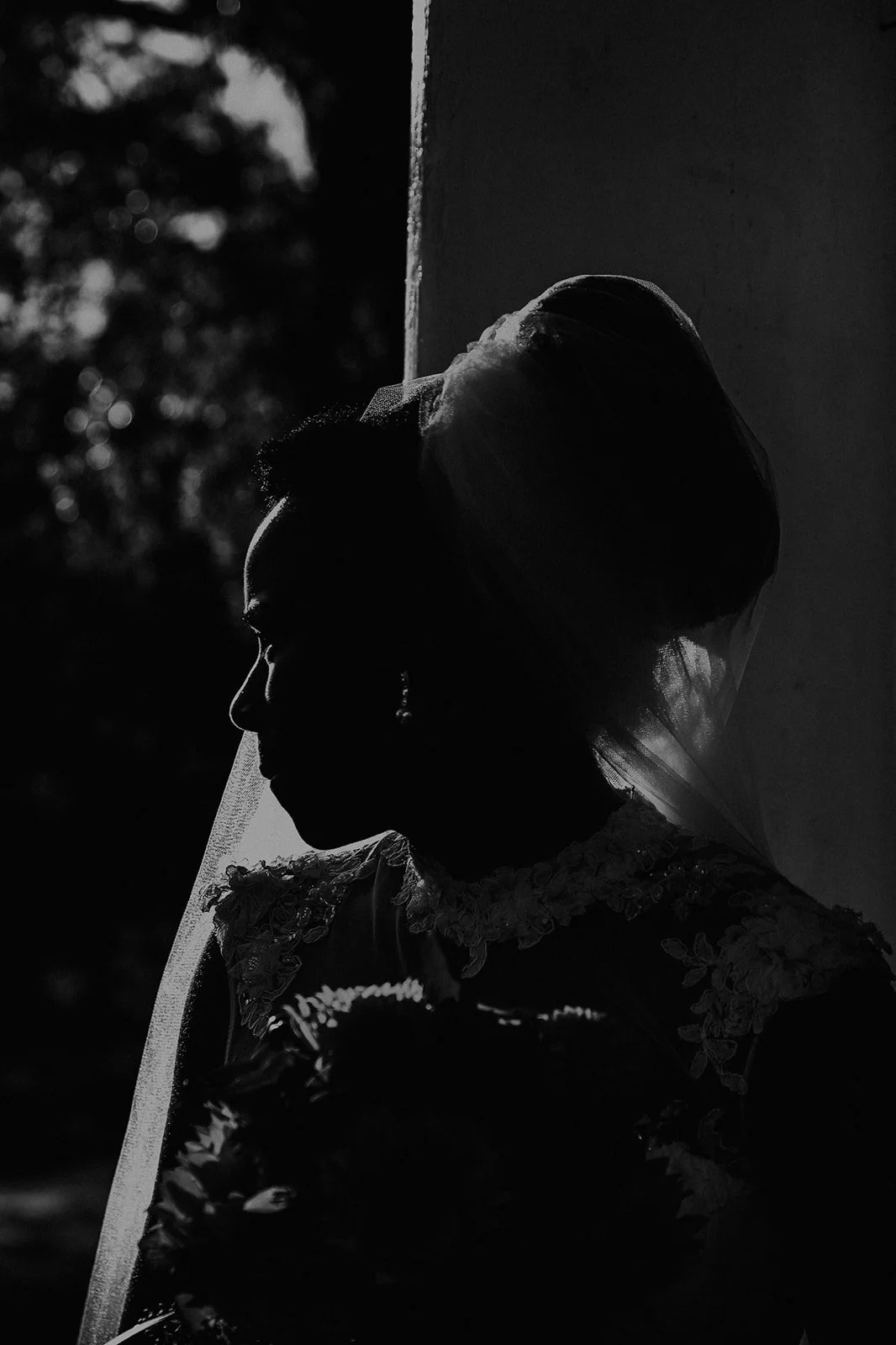 Silhouette of a bride in a wedding dress and veil, holding a bouquet, with dark background and soft lighting highlighting her profile.