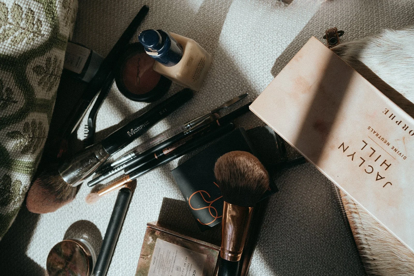 Collection of makeup brushes, a powder compact, a bottle of foundation, and other makeup items on a fabric surface.