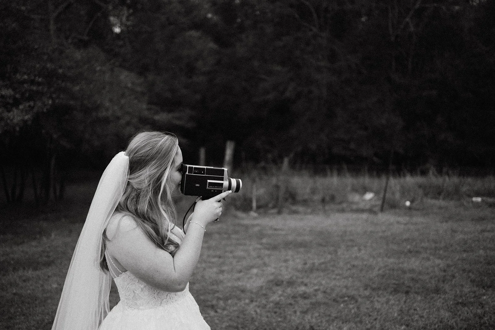 A young woman in a wedding dress and veil is using a vintage Polaroid camera outdoors in a grassy area surrounded by trees.
