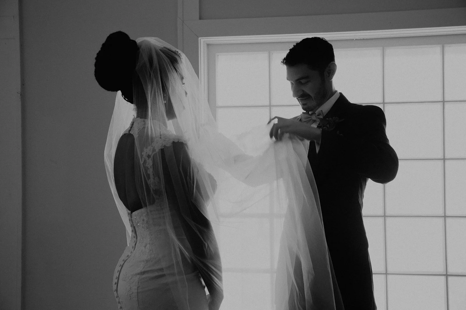 Black and white photo of a groom helping a bride with her veil by a window.