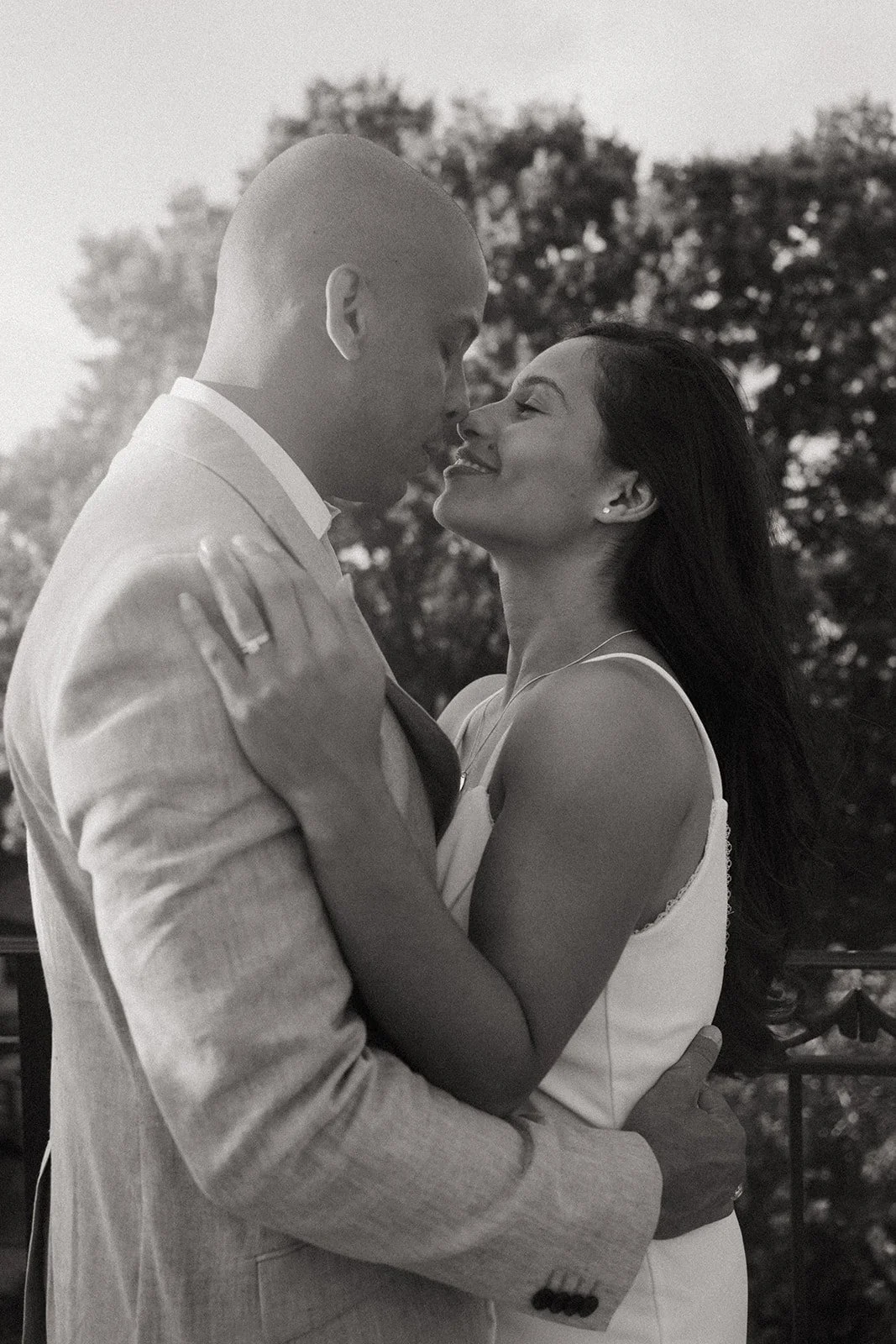 A black-and-white photo of a couple embracing outdoors, about to kiss, with trees in the background during sunset.
