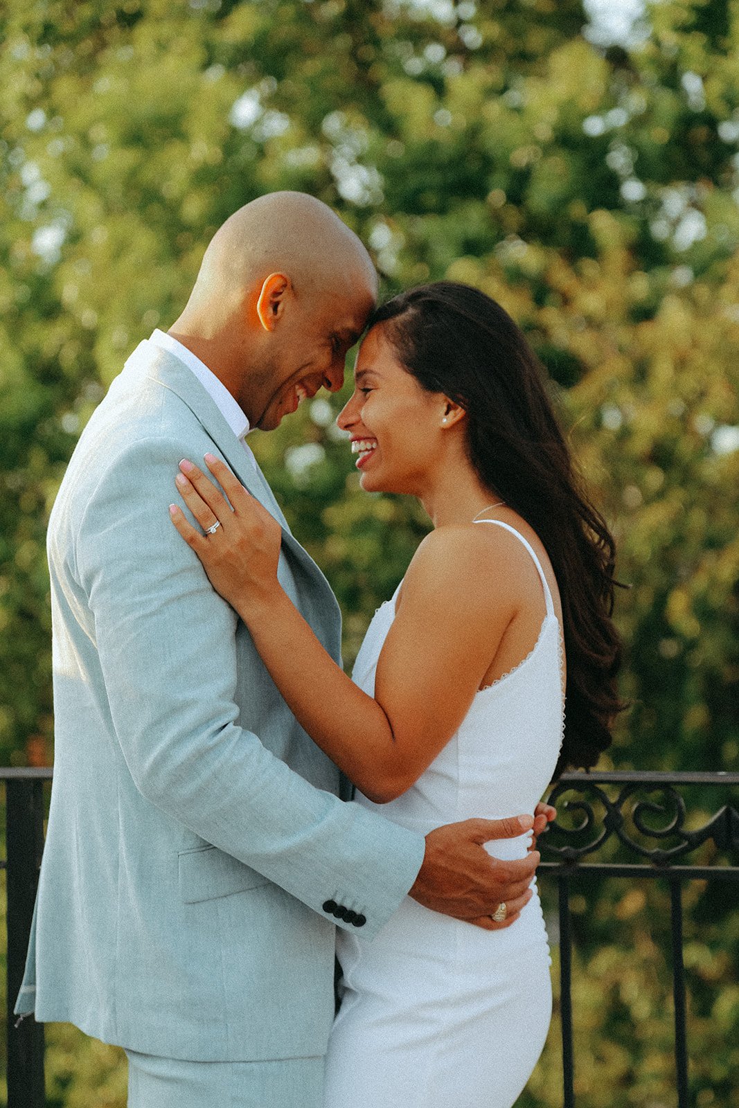 A happy couple is embracing each other outdoors, with trees in the background, smiling and touching foreheads.