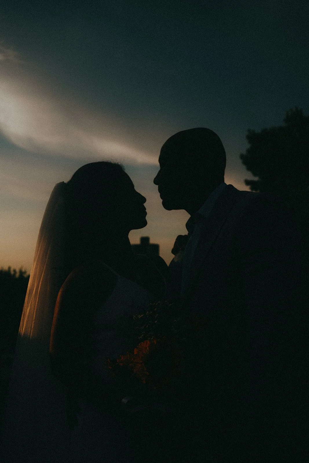 Silhouettes of a couple facing each other outdoors at sunset or dusk, with a dark sky and some clouds in the background.