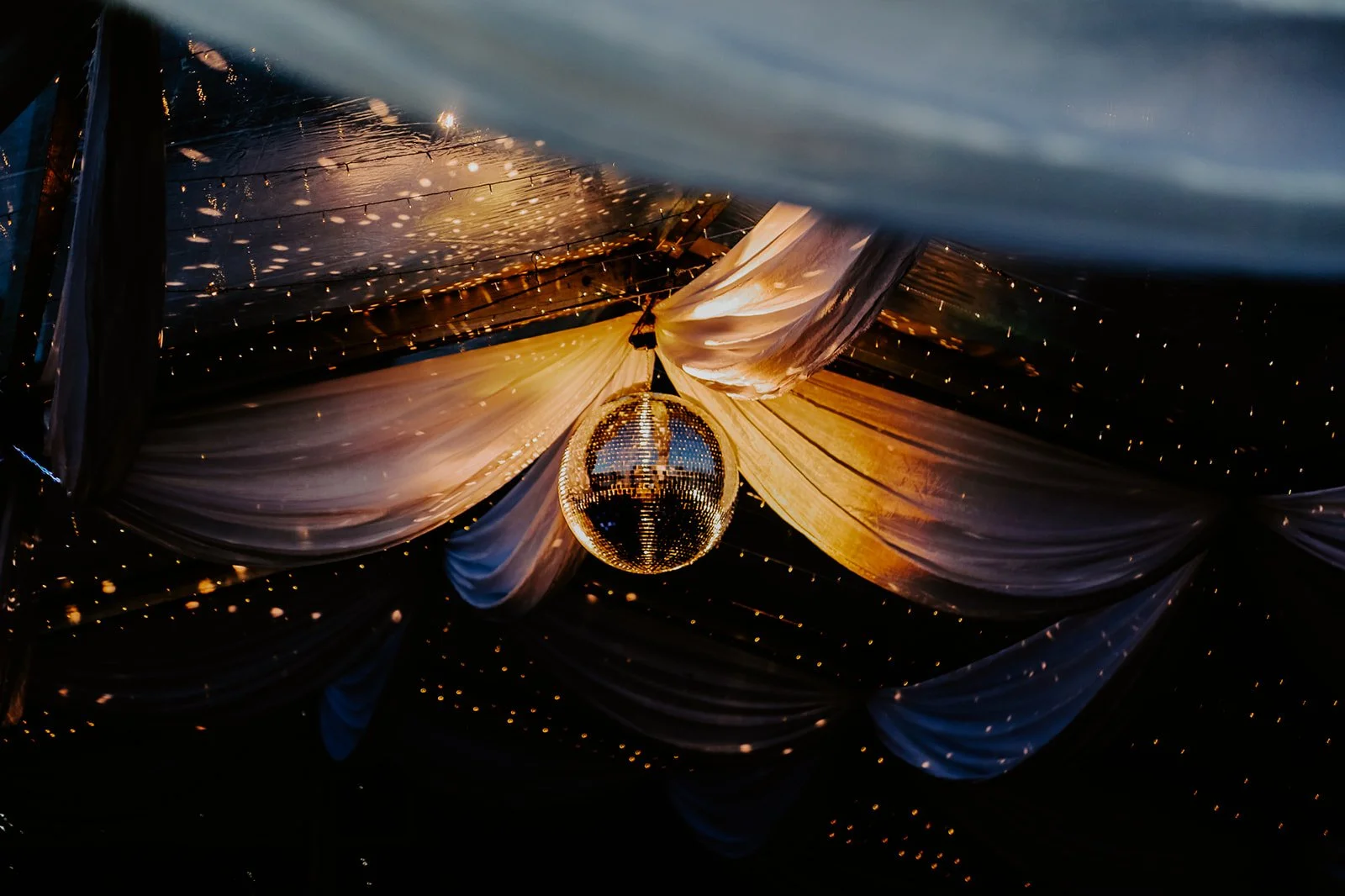 A disco ball hanging from a ceiling decorated with white draped fabric and tiny string lights, creating a festive ambiance.