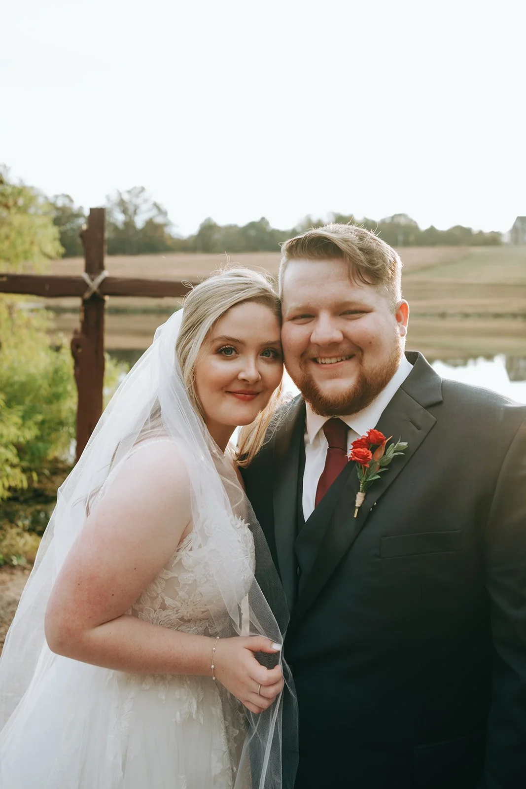 A newlywed couple, a bride with blonde hair and a groom with light brown hair, stands outdoors near a body of water, smiling for the camera on their wedding day.