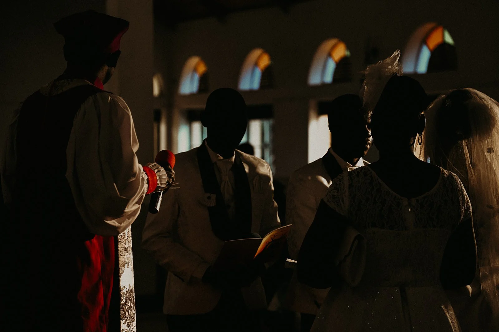 A group of people, dressed in formal attire, standing inside a church or cathedral with arched windows, with some light coming through the windows.