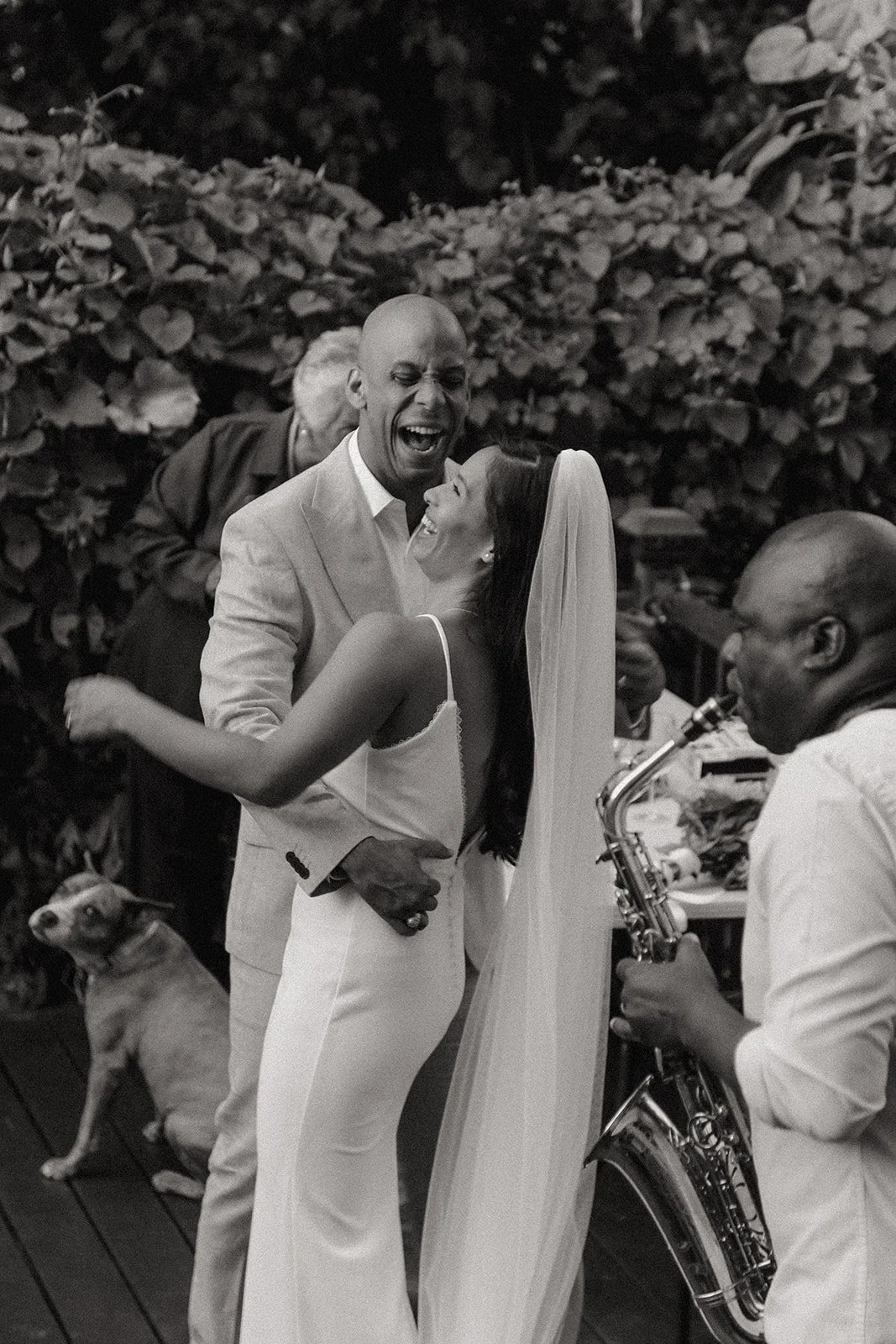 A couple dancing and laughing during a wedding celebration, with a musician playing a saxophone nearby and a dog sitting on the floor.