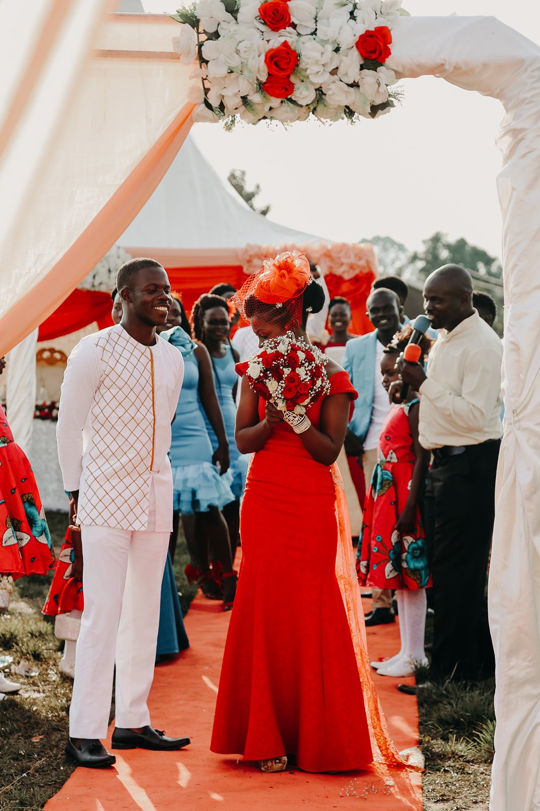 A wedding ceremony with a woman in a red dress holding a bouquet of red and white flowers, standing next to a man in a white traditional outfit. Guests are dressed in bright colors, smiling, and celebrating under a decorated canopy with flowers.