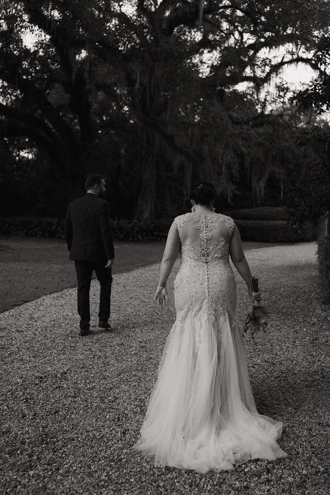A bride in a wedding dress walking away from a groom dressed in a suit along a gravel path in a park or garden with large trees in the background, in black and white.