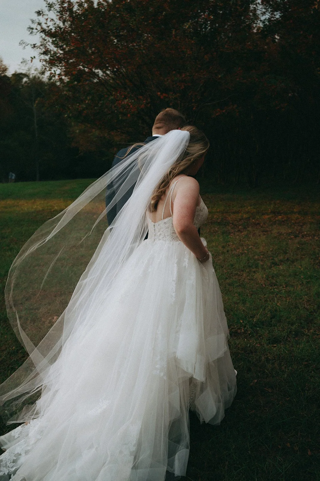 A bride and groom share a kiss outdoors at sunset, with the bride in a white wedding dress and veil and the groom in a dark suit, standing in a grassy field with trees in the background.