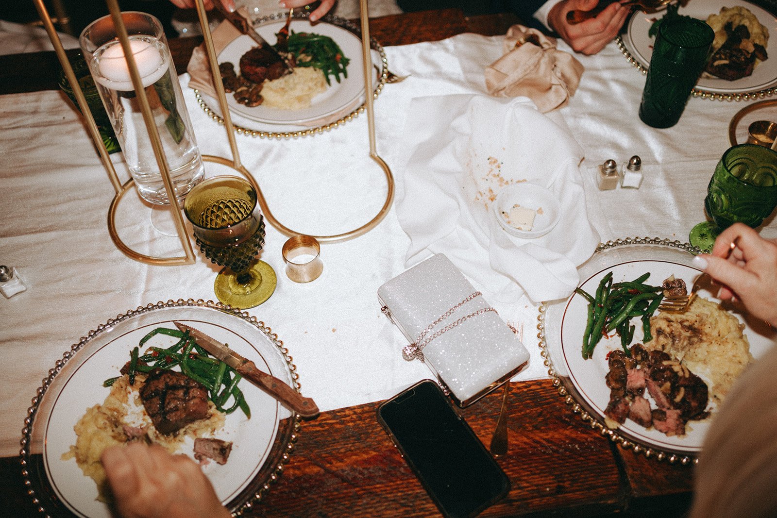 A dining table with plates of food, green glasses, a glass of water, salt and pepper shakers, a sparkly clutch purse, a smartphone, and a decorative gold and glass centerpiece, with people reaching for their plates.